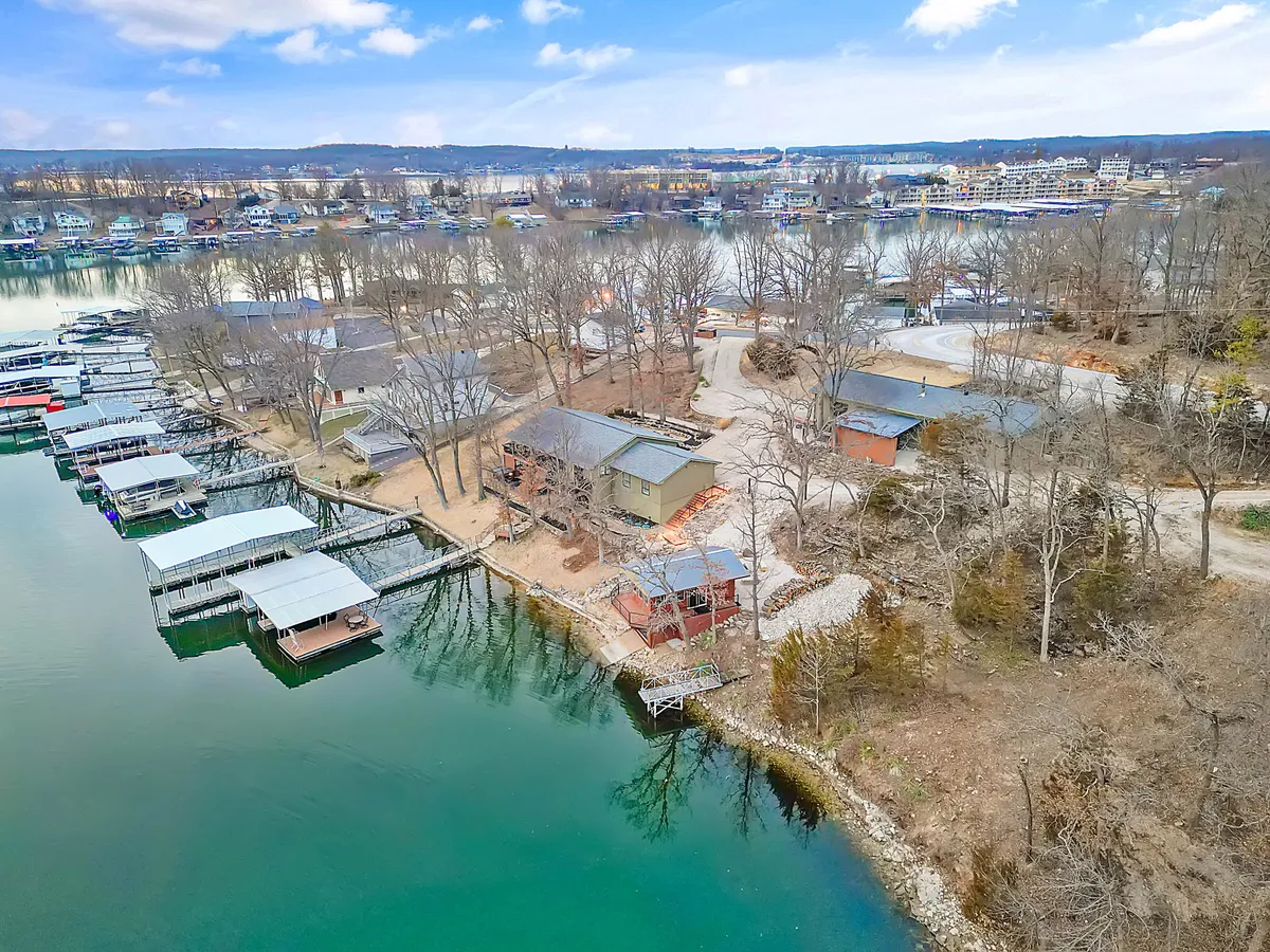 A bustling marina is shown with boats docked in clear water, houses dotting the shoreline, and treed hills in the background. The busy waterfront atmosphere evokes a sense of connection and community, perfect for evenings out or casual group gatherings.