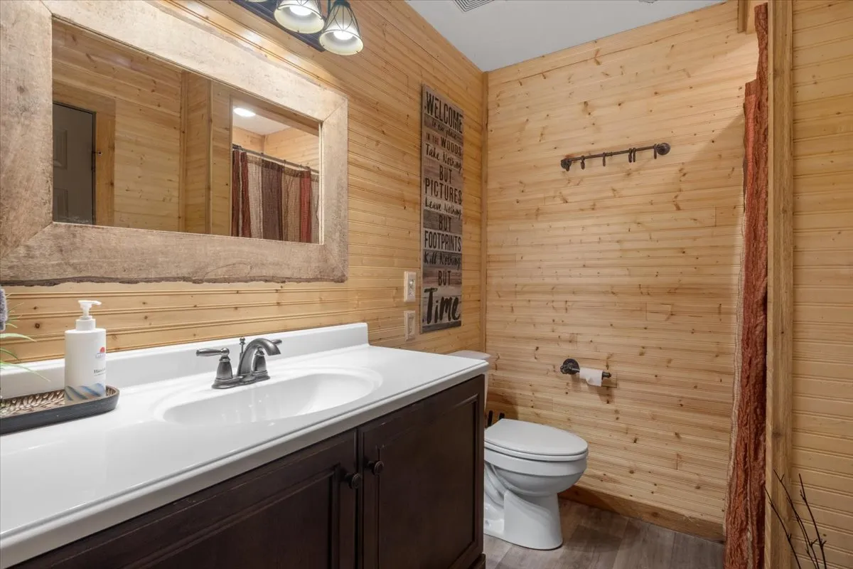 A cozy bathroom scene with a vanity, sink, mirror, wooden walls, and decorative wall art, creating a warm, inviting environment for personal routines and privacy.