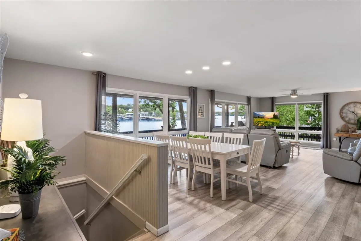 This image captures a welcoming interior with large windows offering panoramic views of the lake, a spacious dining table, and a comfortable seating area adorned with decorative plants. The sunlight floods the space, highlighting the inviting atmosphere ideal for family gatherings, dining, and relaxation.