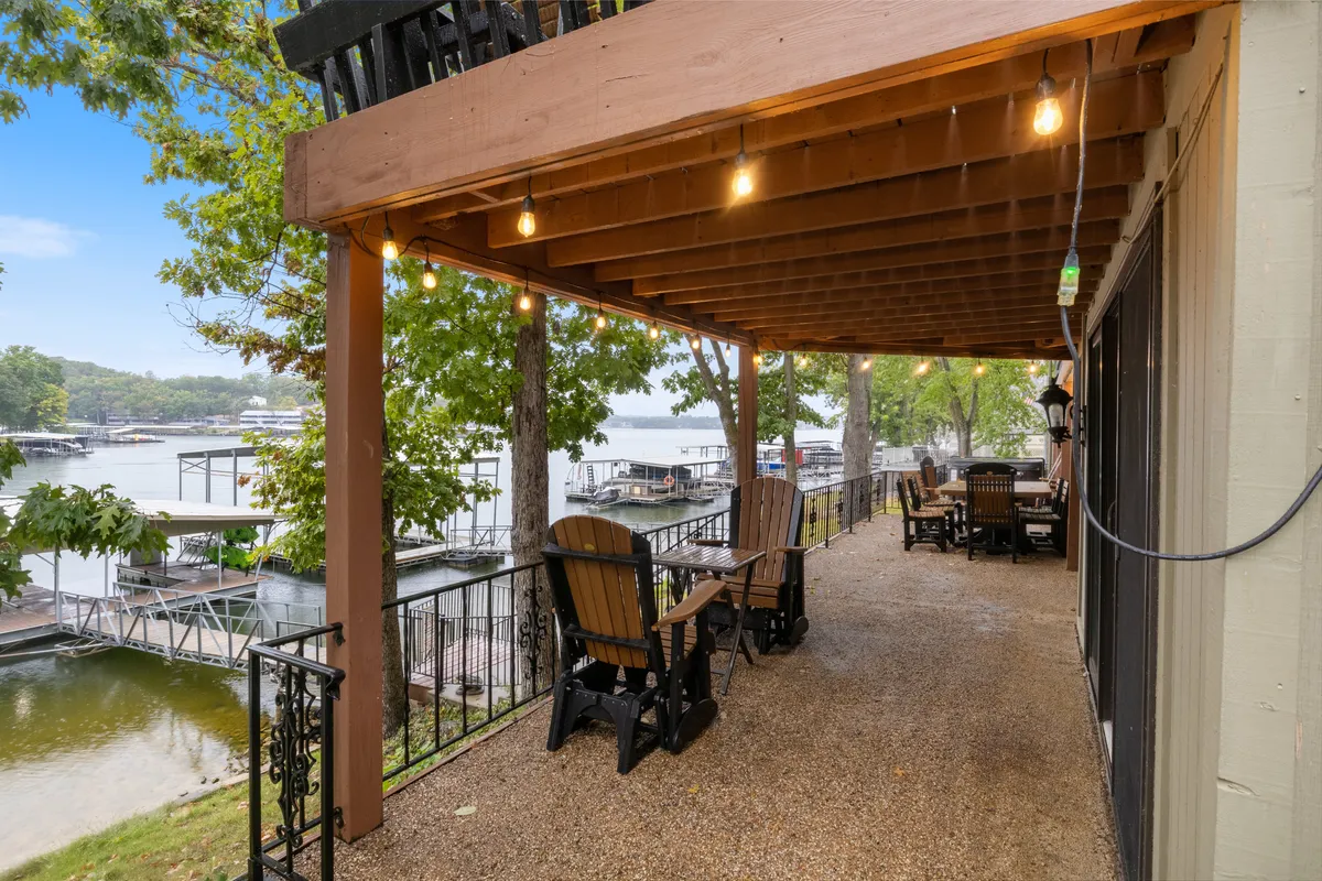 A spacious wooden deck sits beside the lake, featuring comfortable outdoor seating and string lights overhead. The area is designed for gatherings, with a serene waterfront view inviting group relaxation, dining, or socializing. Lush greenery frames the deck, enhancing its tranquil, resort-like ambiance.