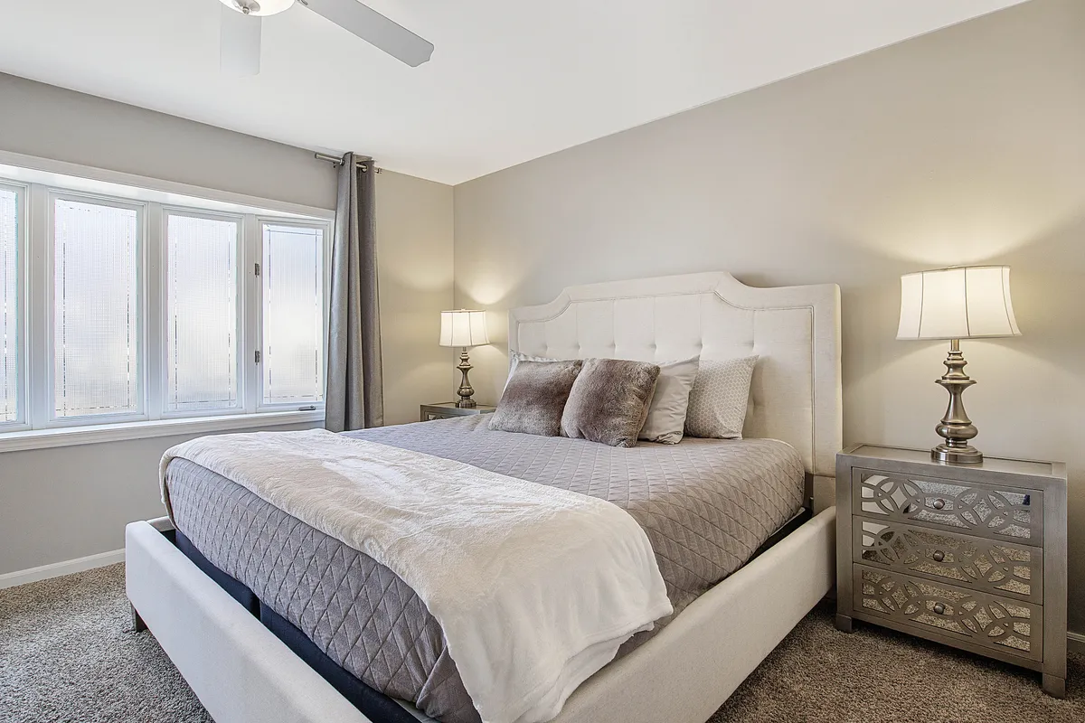 A tranquil bedroom featuring a neatly made bed adorned with decorative pillows, nightstands with lamps on either side, and expansive windows that flood the room with natural light. The design is cozy yet sophisticated, perfect for peaceful slumber.