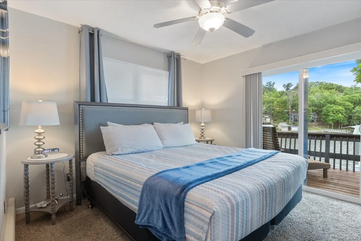 A serene bedroom with blue and white bedding, a large window offering a view of the outdoors, and a decorative bedside lamp. The space radiates comfort and tranquility, ideal for restful moments or reading by natural light.