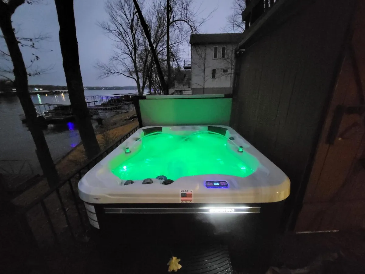 A hot tub illuminated by green lights sits on a deck overlooking the water, surrounded by towering trees, with the lake visible beyond. The scene is tranquil and inviting, perfect for soaking while watching the sun set or stars emerge.