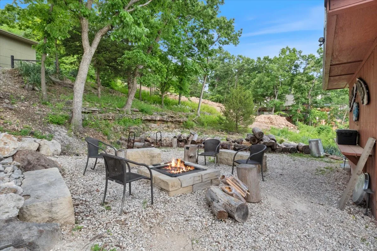 An inviting fire pit area with comfortable seating is set within a leafy green landscape. A stone path winds to the fire pit, designed for gathering around in the evenings for stories, marshmallow roasting, and laughter under the stars.