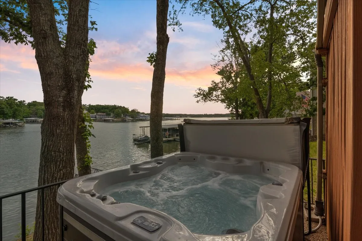 The image showcases a stunning lakefront scene at sunset, with a bubbling hot tub in the foreground surrounded by mature trees. The tranquil waters of Lake Ozark extend into the distance, and warm, golden hues fill the sky, inviting guests to unwind and soak in the view. The setup clearly suggests spaces for relaxing, socializing, and enjoying nature's beauty.