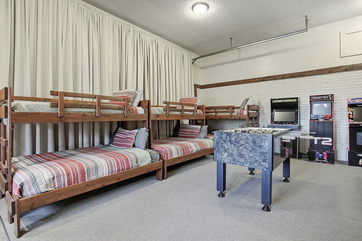 A vibrant interior scene showing multiple twin-over-full bunk beds, a foosball table, and classic arcade game machines. The space is brightly lit and designed for both play and rest, with ample room for social activities and relaxation.