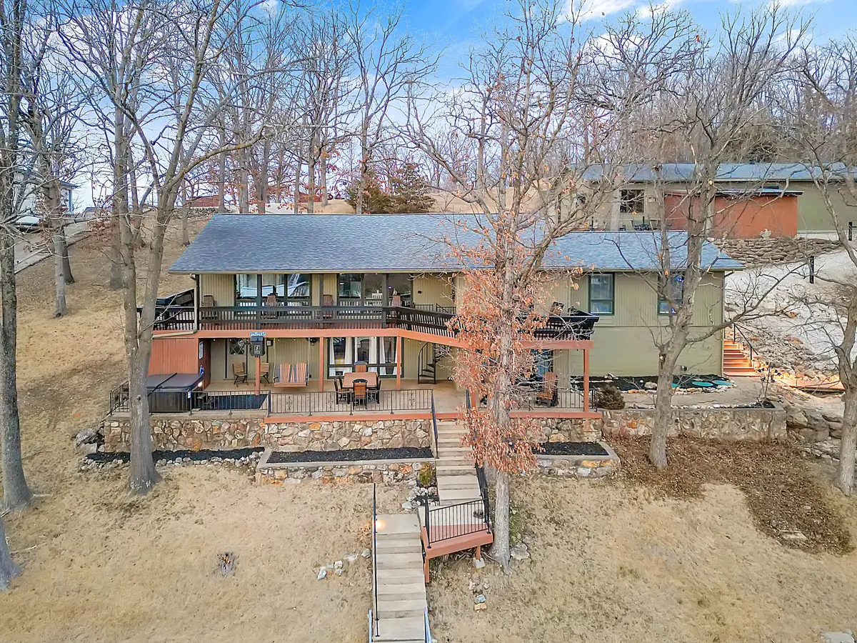 A two-story house framed by trees, with a welcoming deck and stairs leading up to the home. The exterior view showcases the main house, its ample outdoor space, and the lush natural setting, suggesting plenty of room for large groups and easy access to the outdoors.