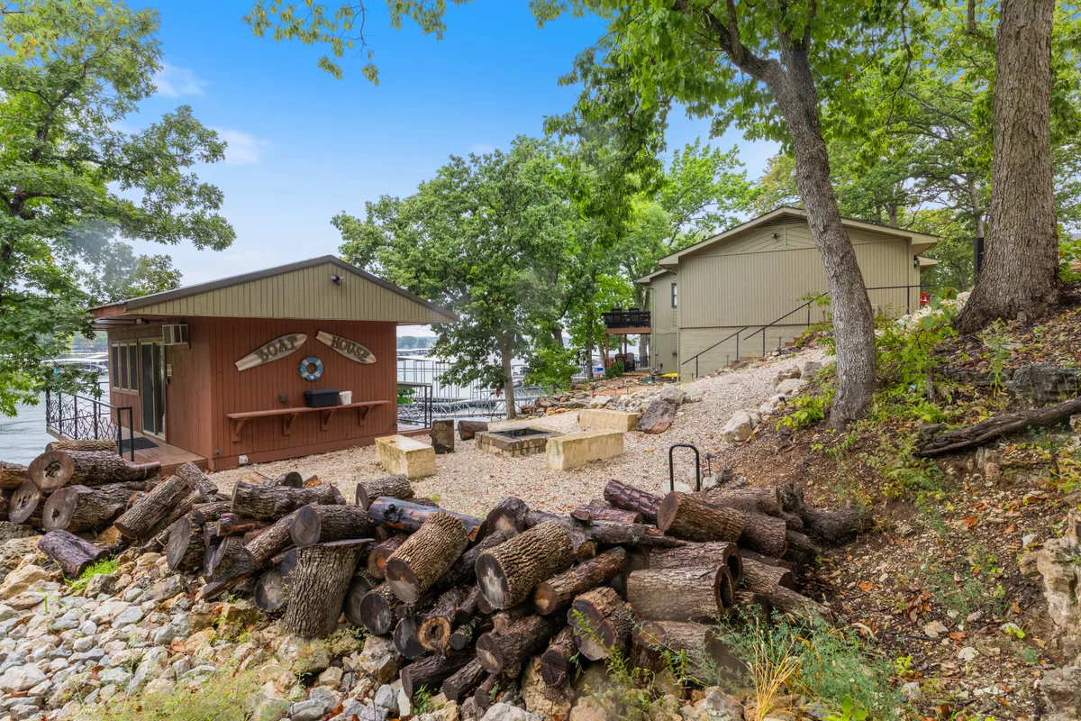An inviting boathouse structure sits near a stone fire pit with stacked logs, shaded by trees and overlooking the water. The scene is set for relaxing by the fire after a day on the lake, with a sense of comfort and exclusivity.